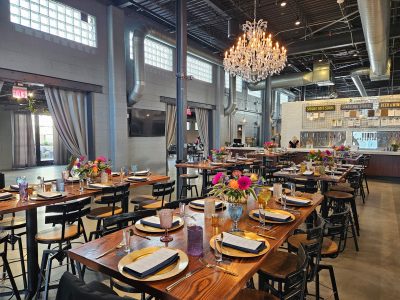 Wedding reception setup at Karrikin Spirits featuring long wood tables, colorful floral centerpieces, crystal chandeliers, and industrial-modern decor.